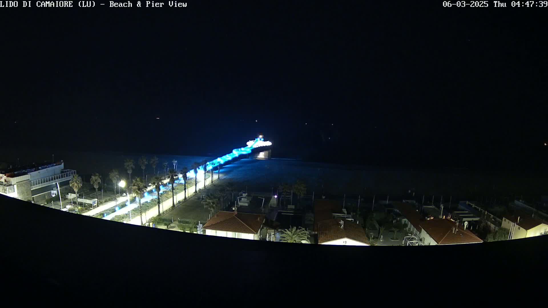 A nighttime, high-angle view shows a pier illuminated in blue light extending from a shoreline into dark water, with buildings and palm trees visible along the beach.