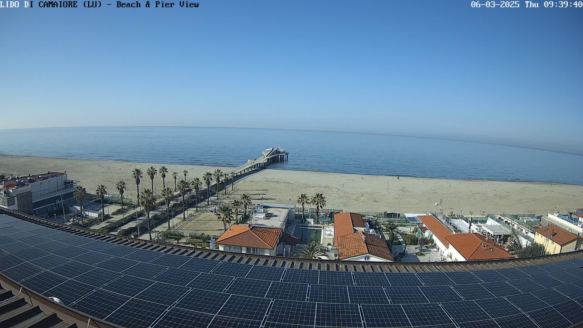 A long pier extends from a sandy beach into a calm ocean under a clear blue sky, viewed from a rooftop covered in solar panels.