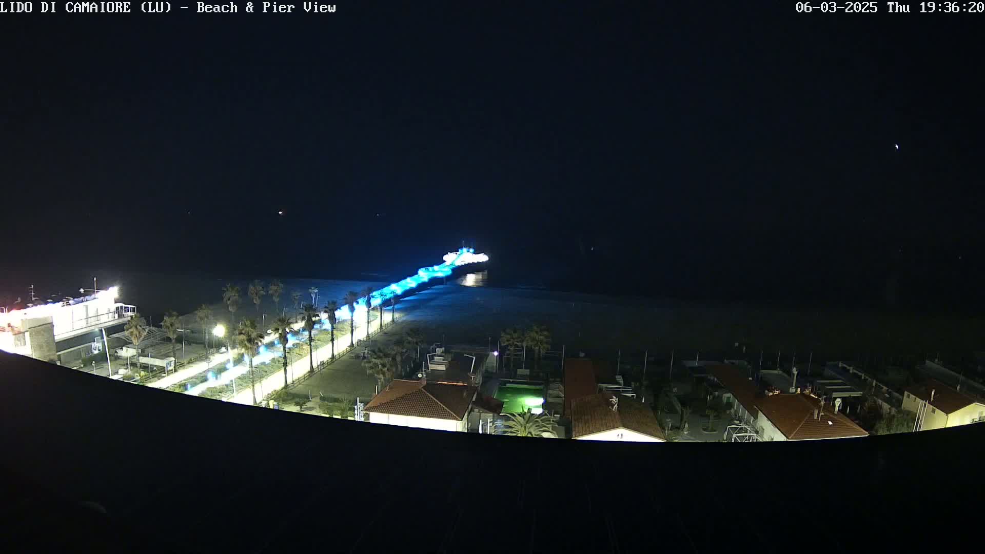 A nighttime, high-angle view shows a beach and pier illuminated with artificial lights, with calm water and a few palm trees visible along the shore.
