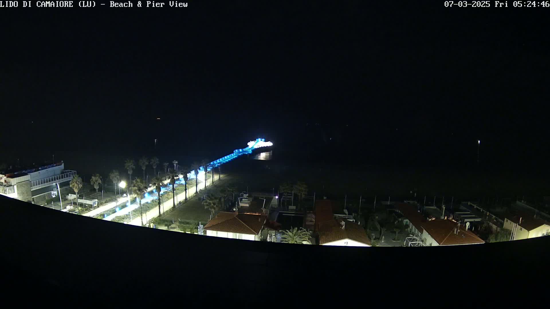 A nighttime, high-angle view shows a beach, pier illuminated with blue lights, and buildings along the shoreline.