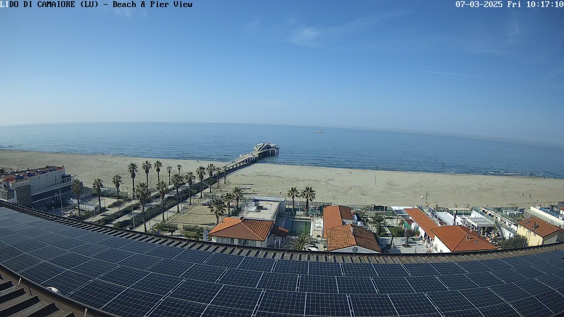A beach with a pier extends into a calm blue ocean under a clear blue sky, viewed from above a rooftop covered in solar panels.