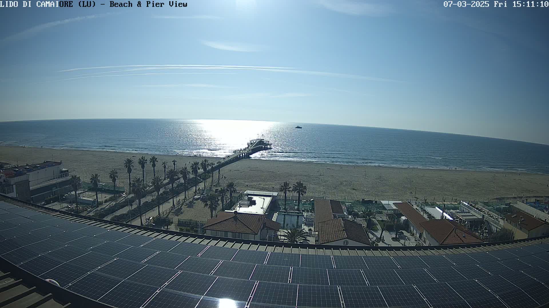 A sandy beach with a pier and palm trees is visible from a rooftop covered in solar panels, under a clear sunny sky.