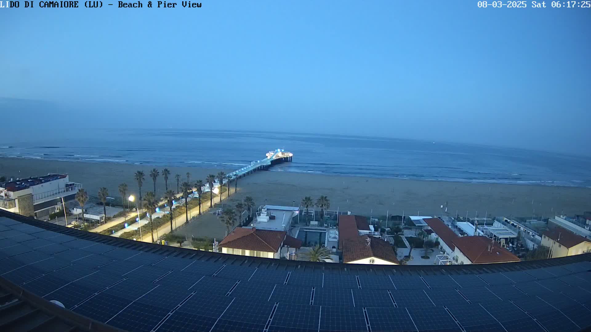 A beach with a pier extends into a calm, twilight-blue ocean, viewed from a rooftop covered in solar panels.
