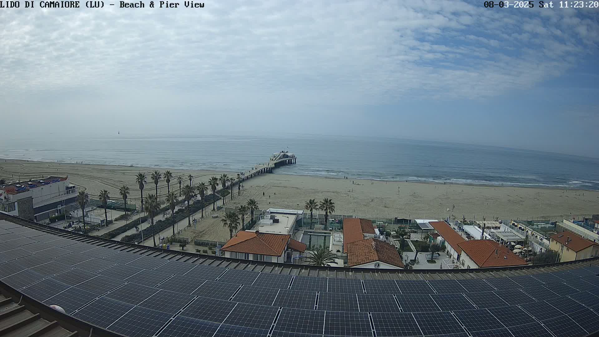 A sandy beach with a pier extends into a calm ocean under a partly cloudy sky, viewed from above a rooftop covered in solar panels.