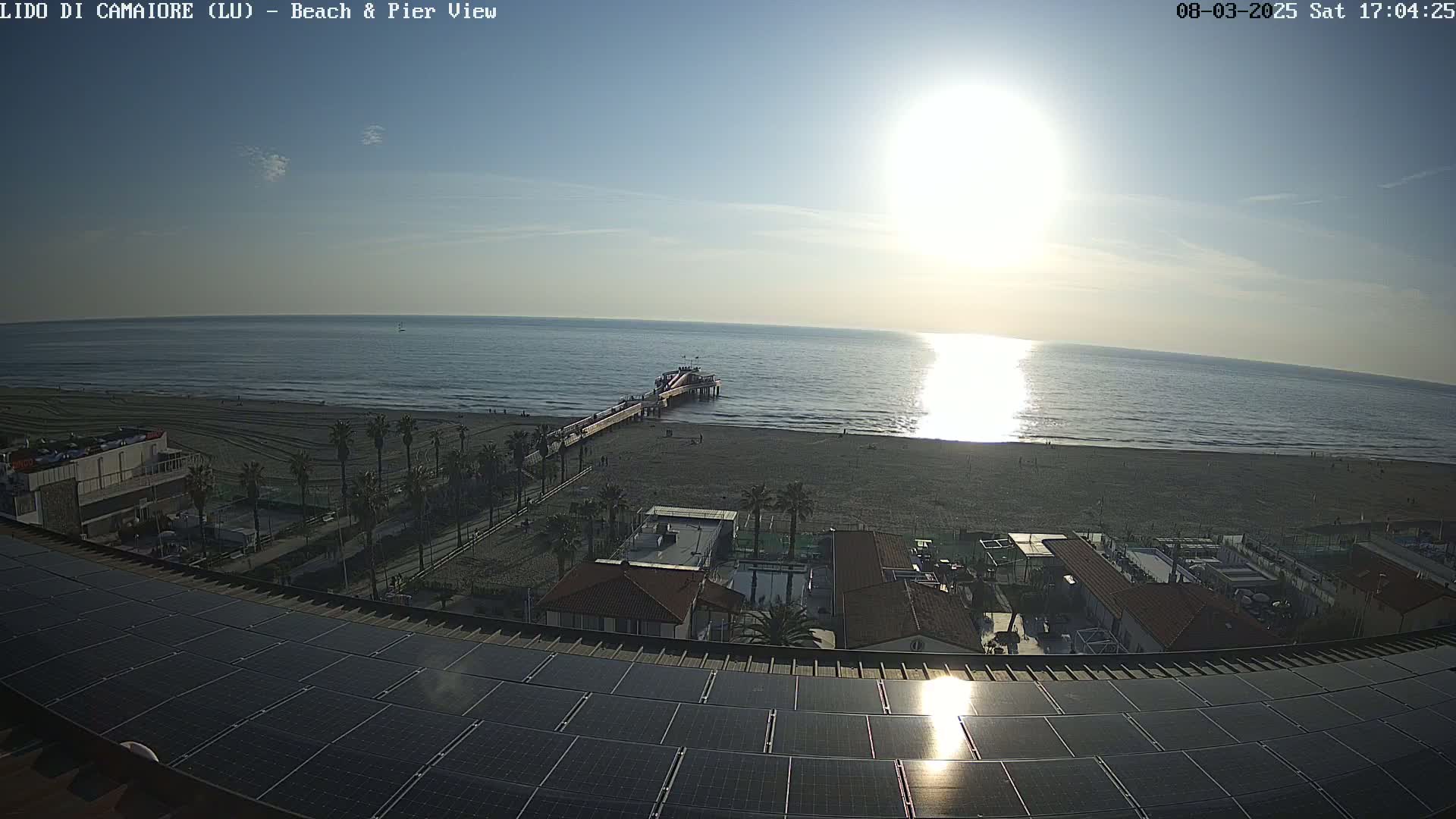 A beach scene with a pier extending into a calm sea under a bright, sunny sky is viewed from a rooftop covered in solar panels.