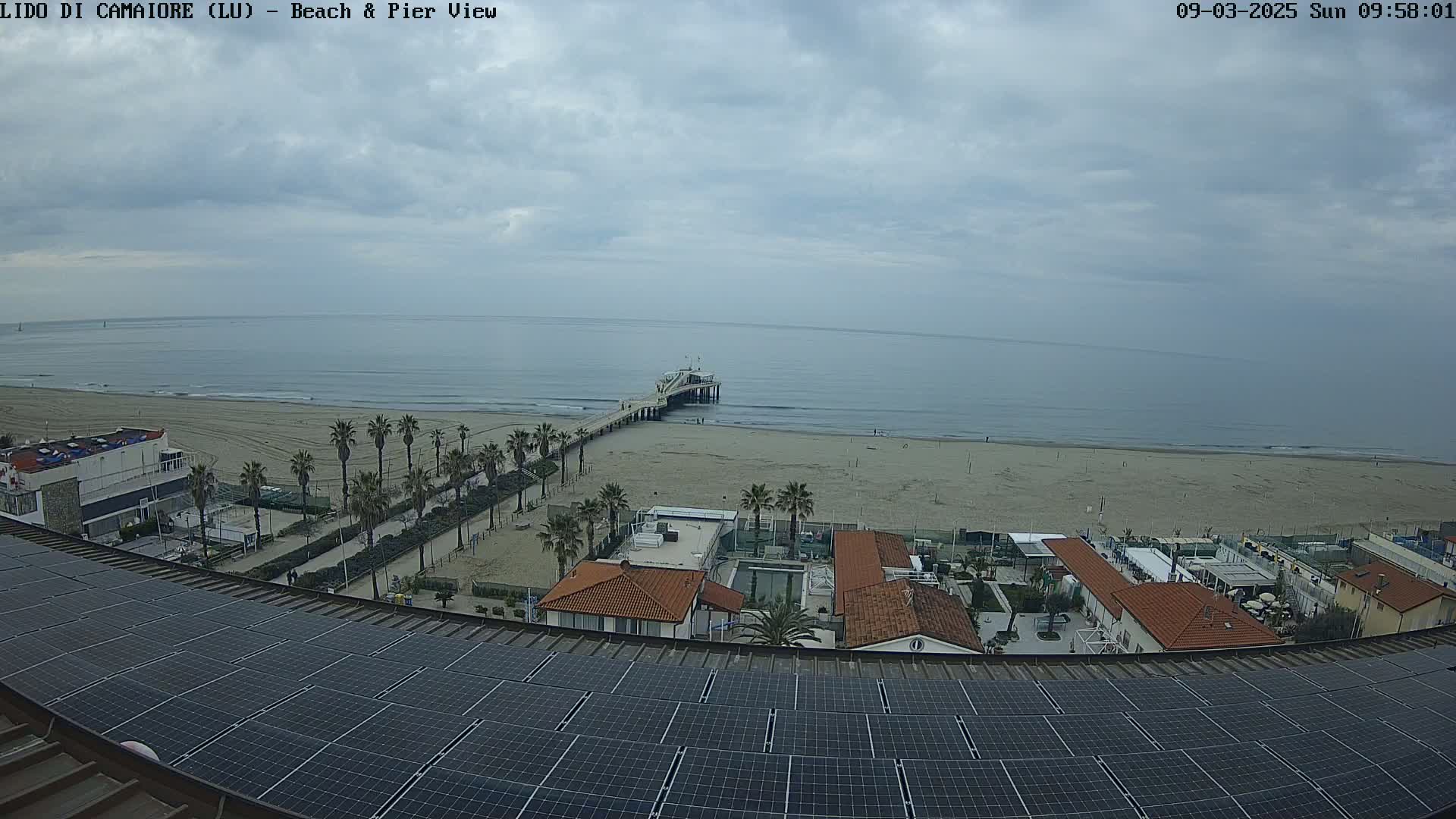 A beach with a pier extends into a calm sea under a cloudy sky, viewed from above a building topped with solar panels.