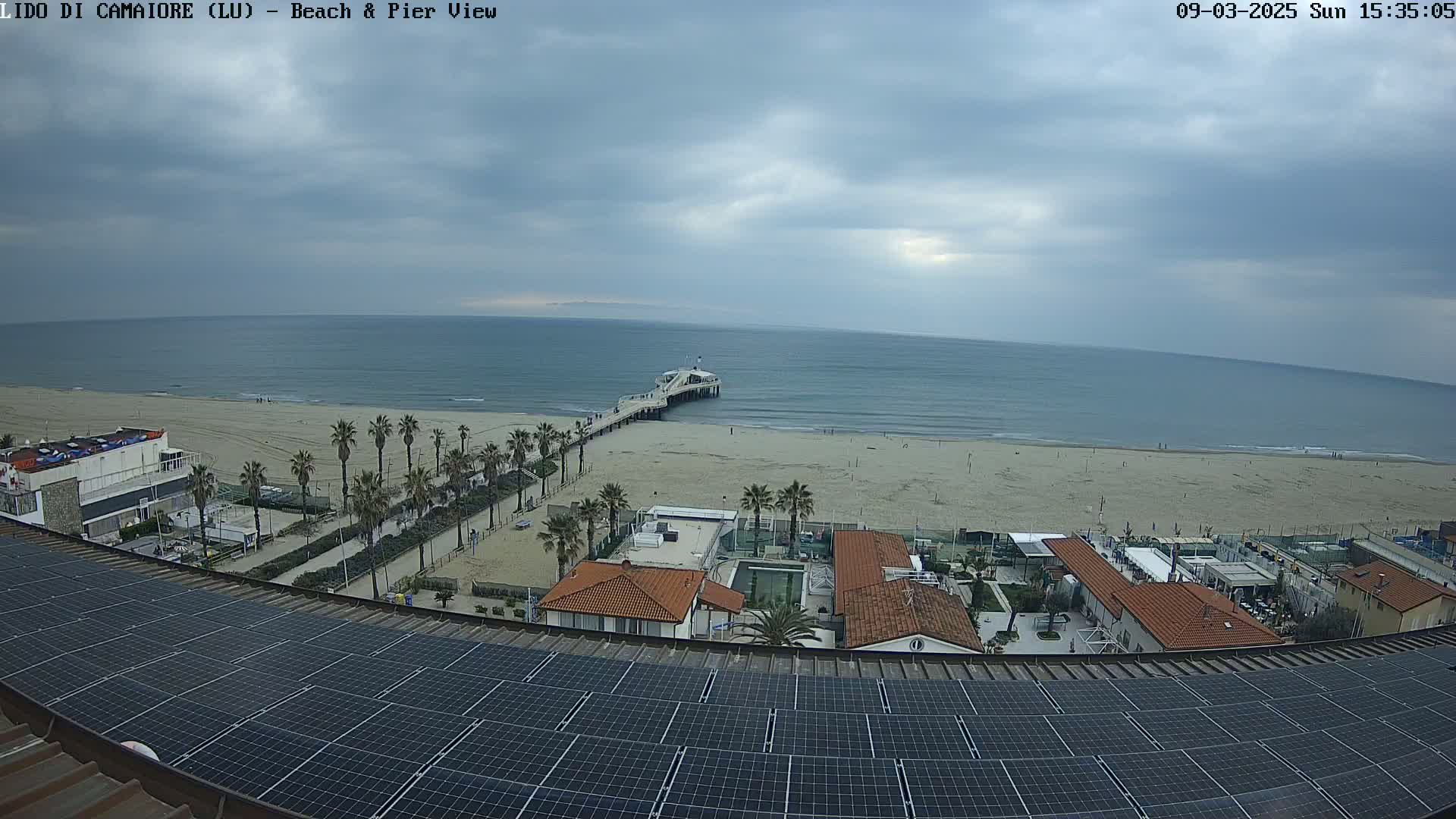 A beach with a pier, palm trees, and buildings is viewed from a rooftop covered in solar panels under a cloudy sky.