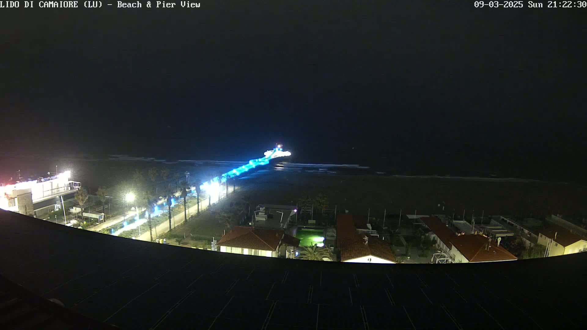 A nighttime, high-angle view shows a pier with blue lights extending into a dark ocean under a mostly clear sky, with a beach and buildings visible below.