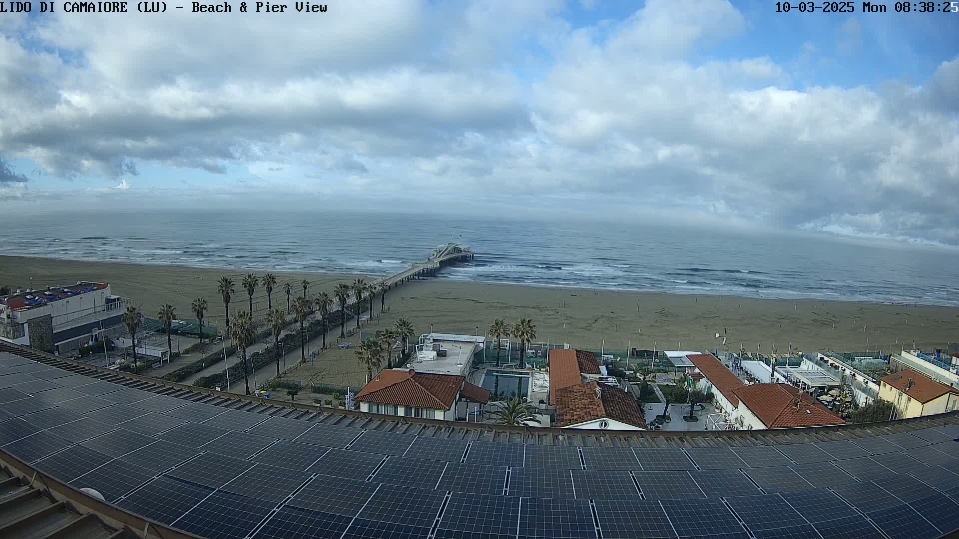 A beach with a pier and several buildings is viewed from above on a partly cloudy day, with a large solar panel array in the foreground.