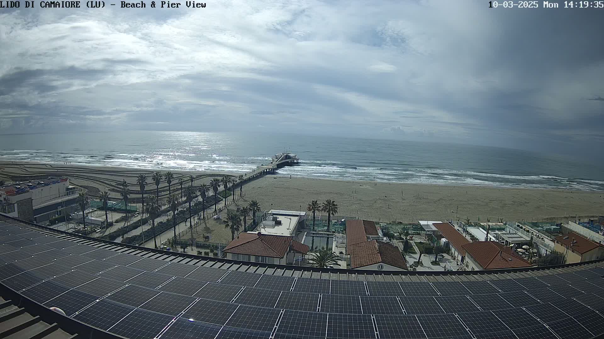 A beach with a pier and buildings is seen from an elevated vantage point under a mostly cloudy sky.