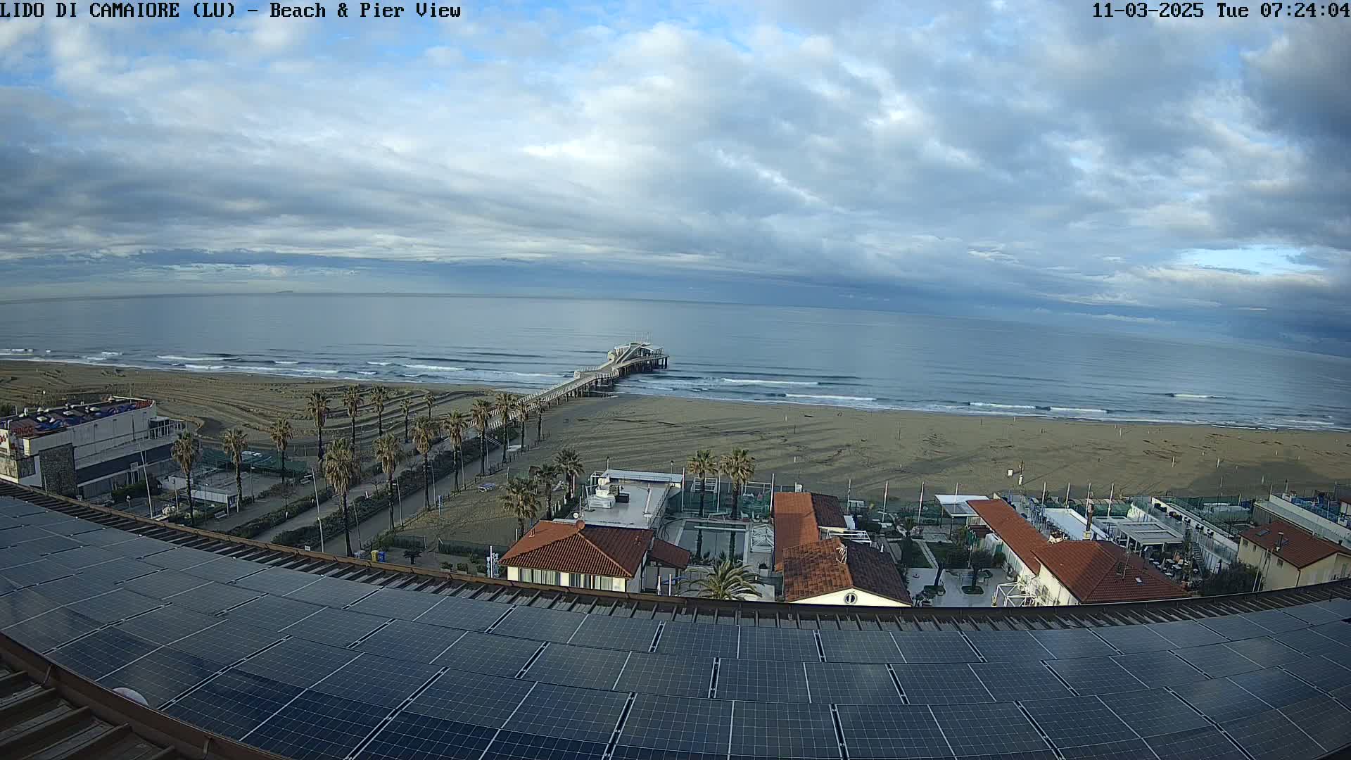 A beach with a pier extends into a wavy ocean under a partly cloudy sky, viewed from a rooftop with solar panels.