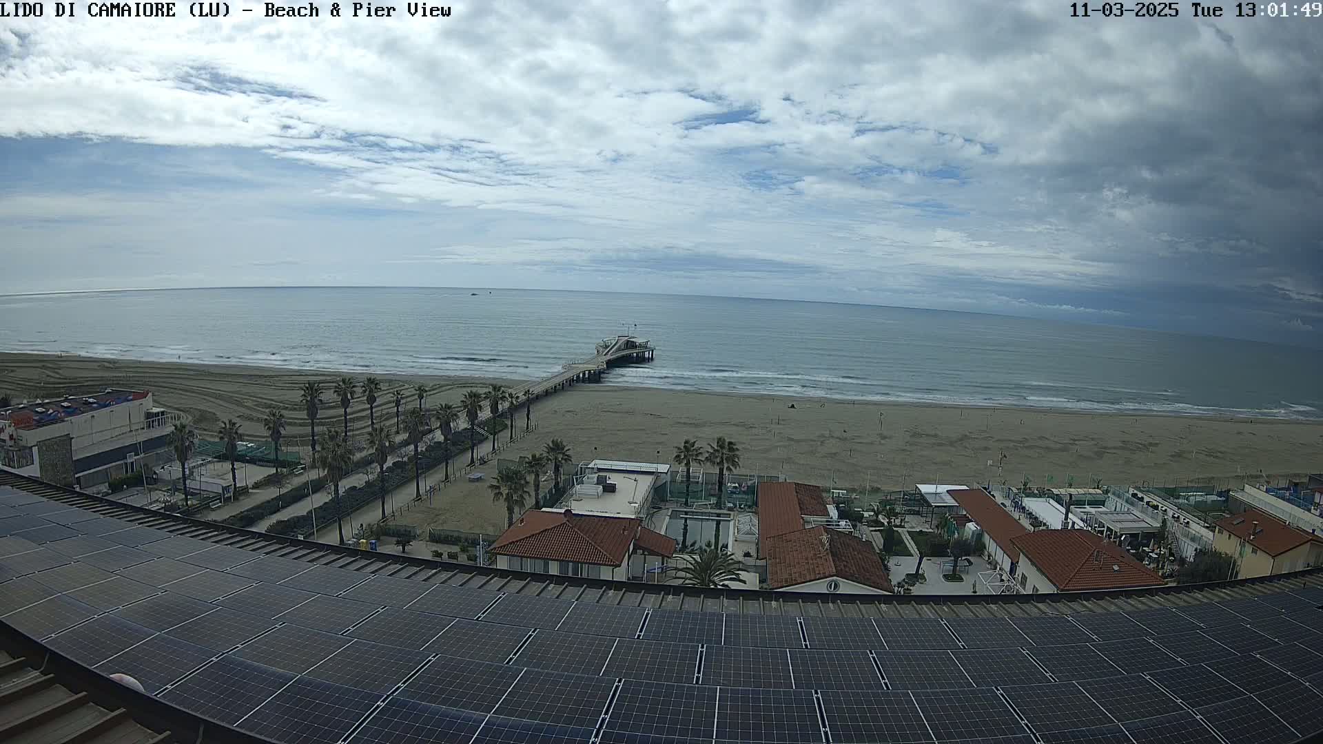 A mostly cloudy day overlooking a sandy beach, a pier extending into the ocean, and a rooftop covered in solar panels with buildings and palm trees in the foreground.