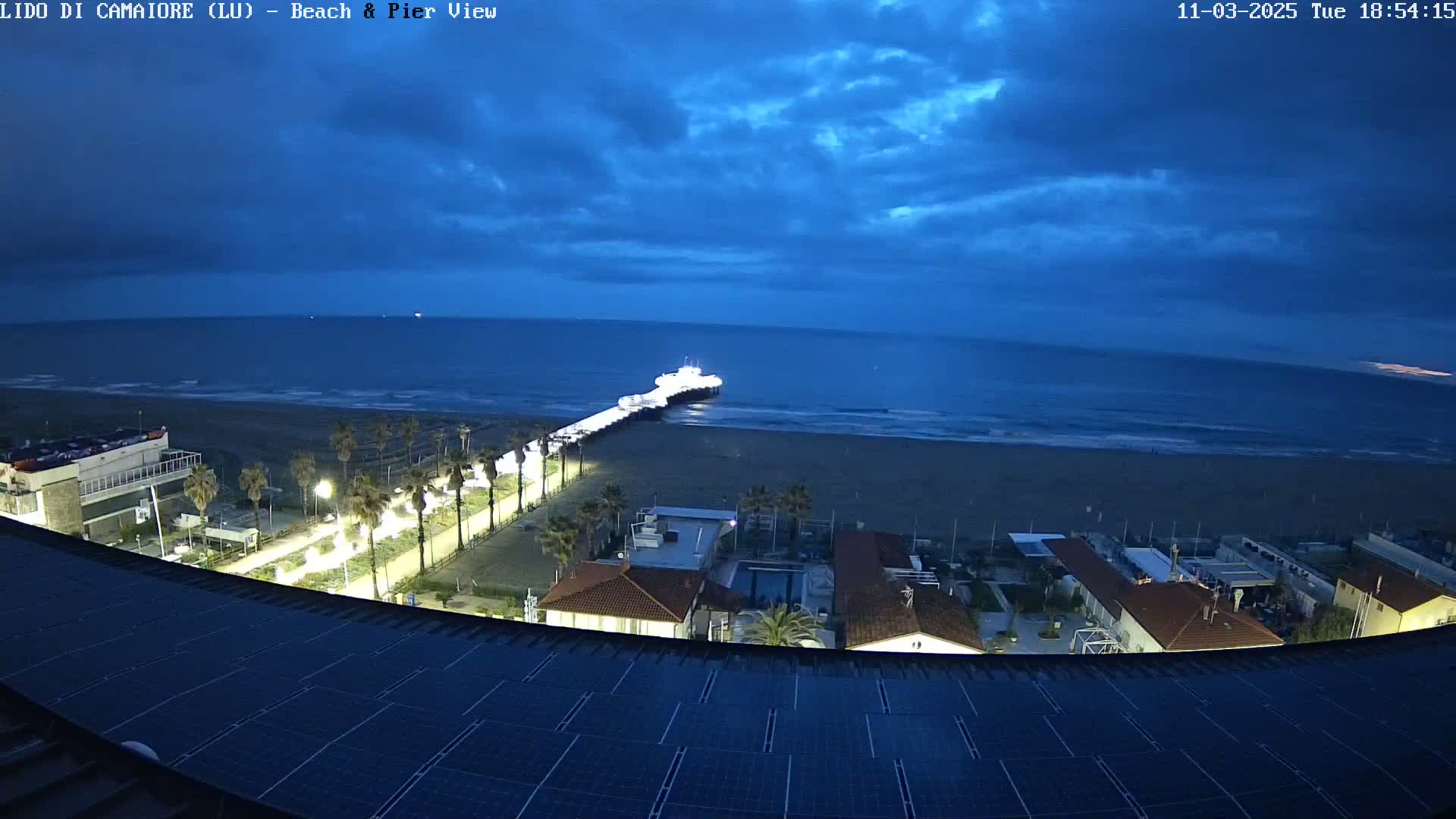 A nighttime view of a beach, pier, and coastal buildings under a partly cloudy sky.