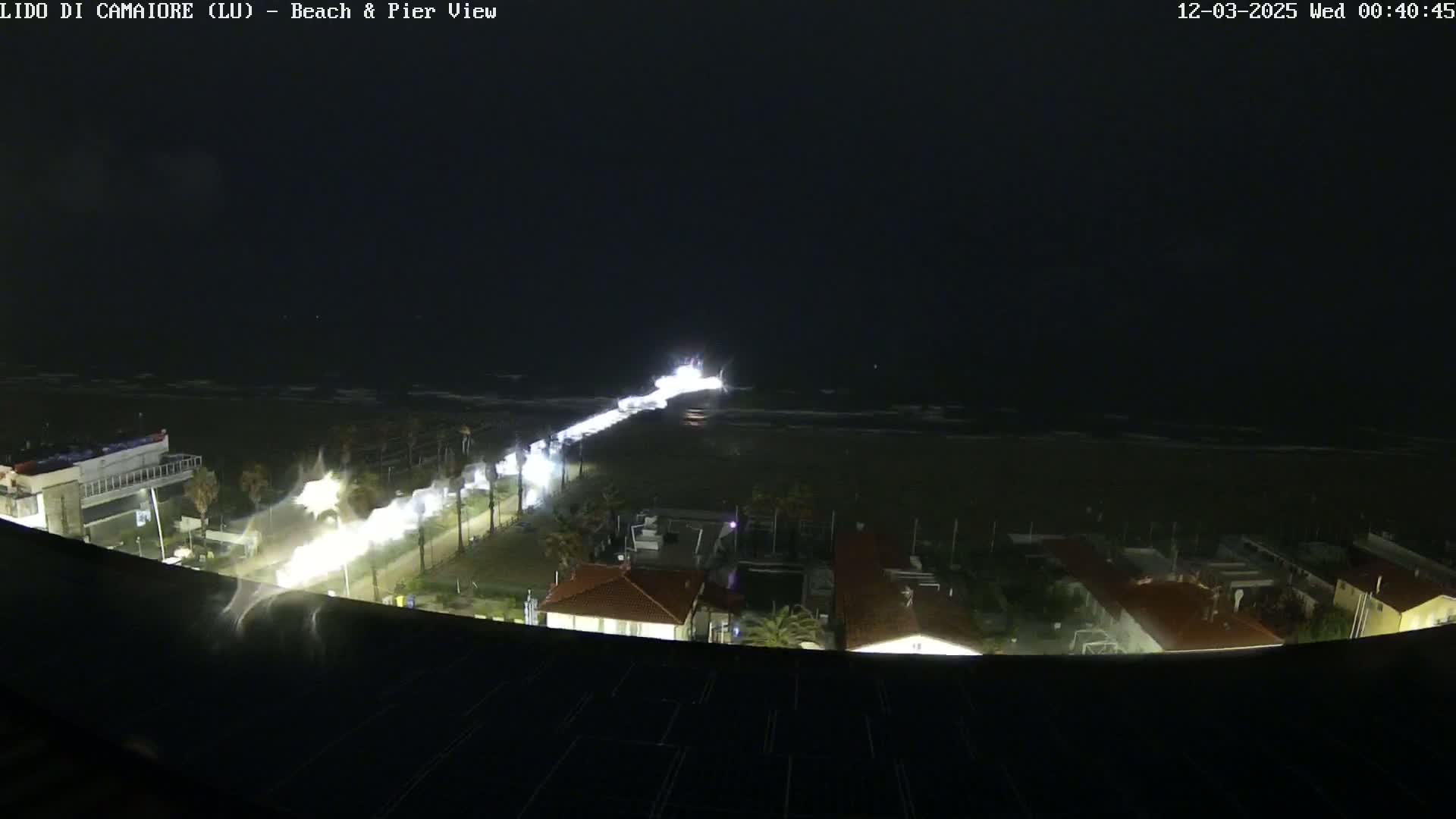 A nighttime, high-angle view of a beach and coastline showing a dark ocean with waves, a brightly lit road leading to the beach, and buildings along the coast under dark, cloudy skies.