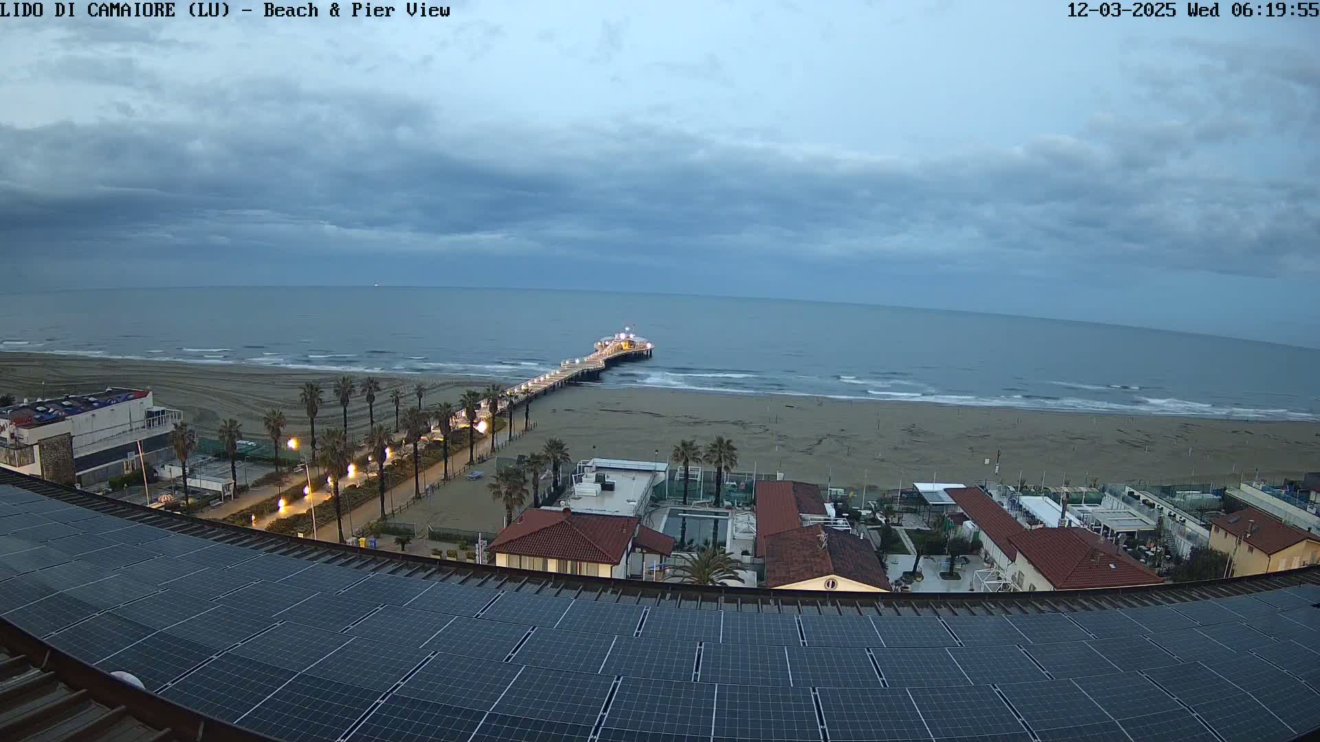 A long pier extends into a calm sea under a cloudy, dusky sky, overlooking a sandy beach and beachfront buildings with a solar panel array in the foreground.