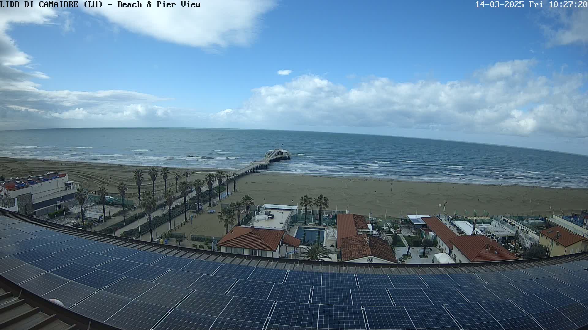 A beach with a pier, palm trees, buildings, and a solar panel array on a rooftop are visible under a partly cloudy sky.