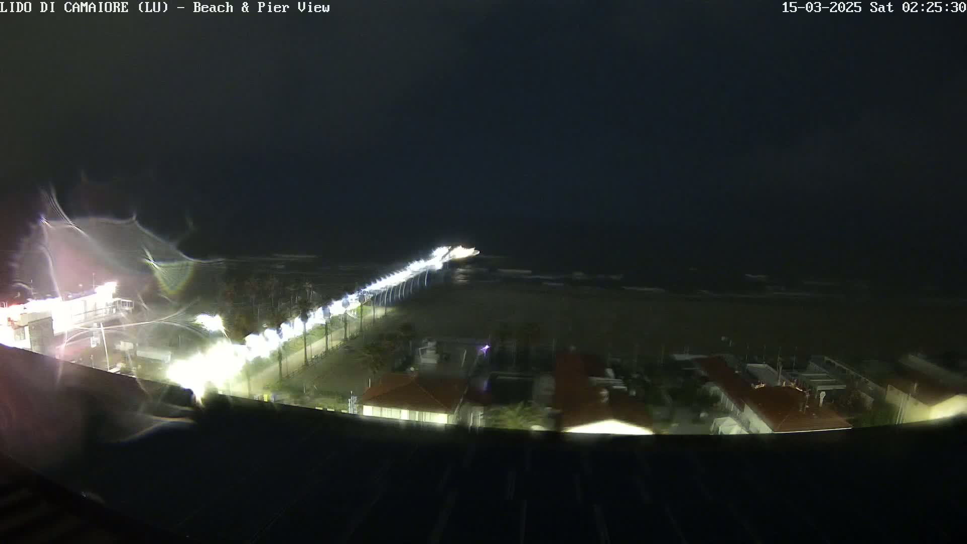 A nighttime view of a beach and pier with rough seas under dark, cloudy skies.