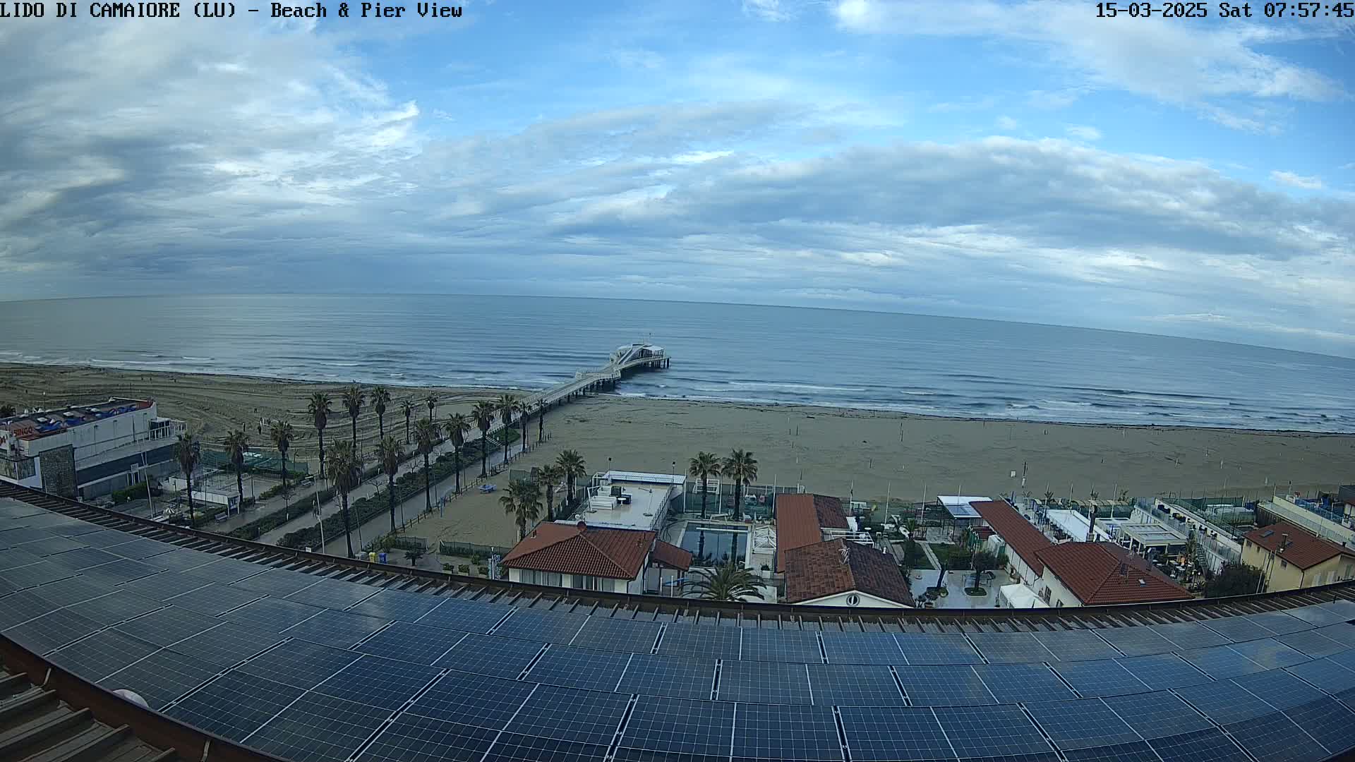 A view from a rooftop with solar panels shows a beach with a pier extending into a somewhat cloudy ocean.