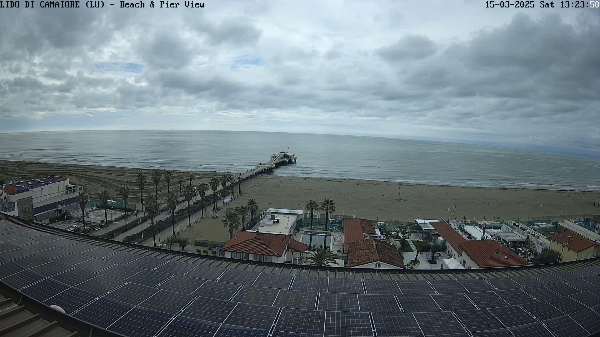 A beach scene under a cloudy sky shows a pier extending into a calm sea, palm trees lining a sandy shore, and buildings with solar panels on a rooftop.