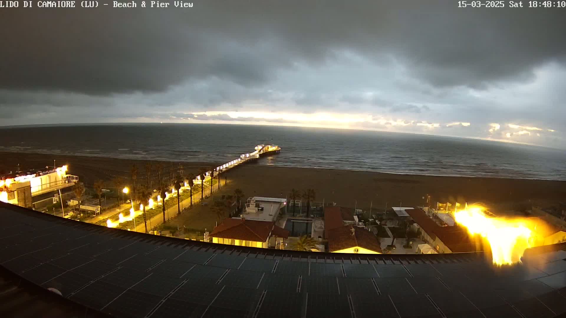 A long pier extends from a beach into a dark, overcast ocean at sunset, with illuminated palm trees and buildings visible along the shoreline, and a large fire or bright light appearing near the lower right.