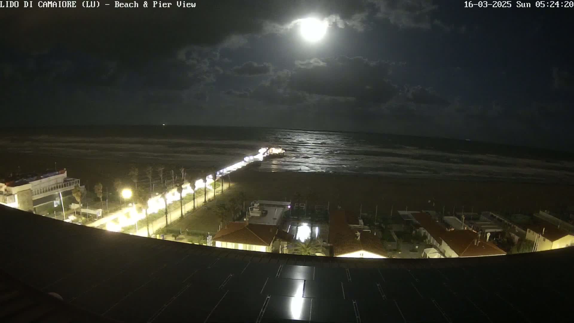 A nighttime view of a beach and pier under a bright moon with some clouds in the sky, showing a coastal town with illuminated buildings and roads.