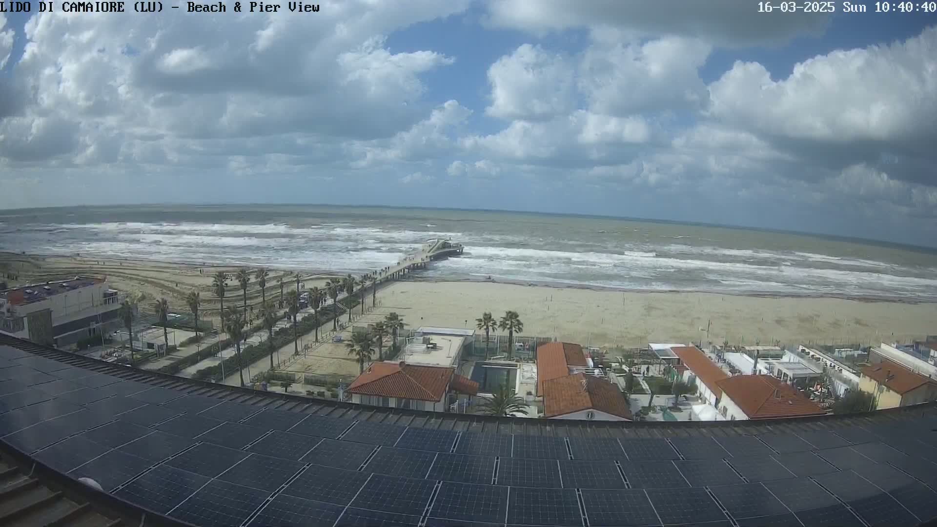 A partly cloudy day shows a beach with a pier, waves crashing on the shore, and a row of buildings with solar panels on the roof.