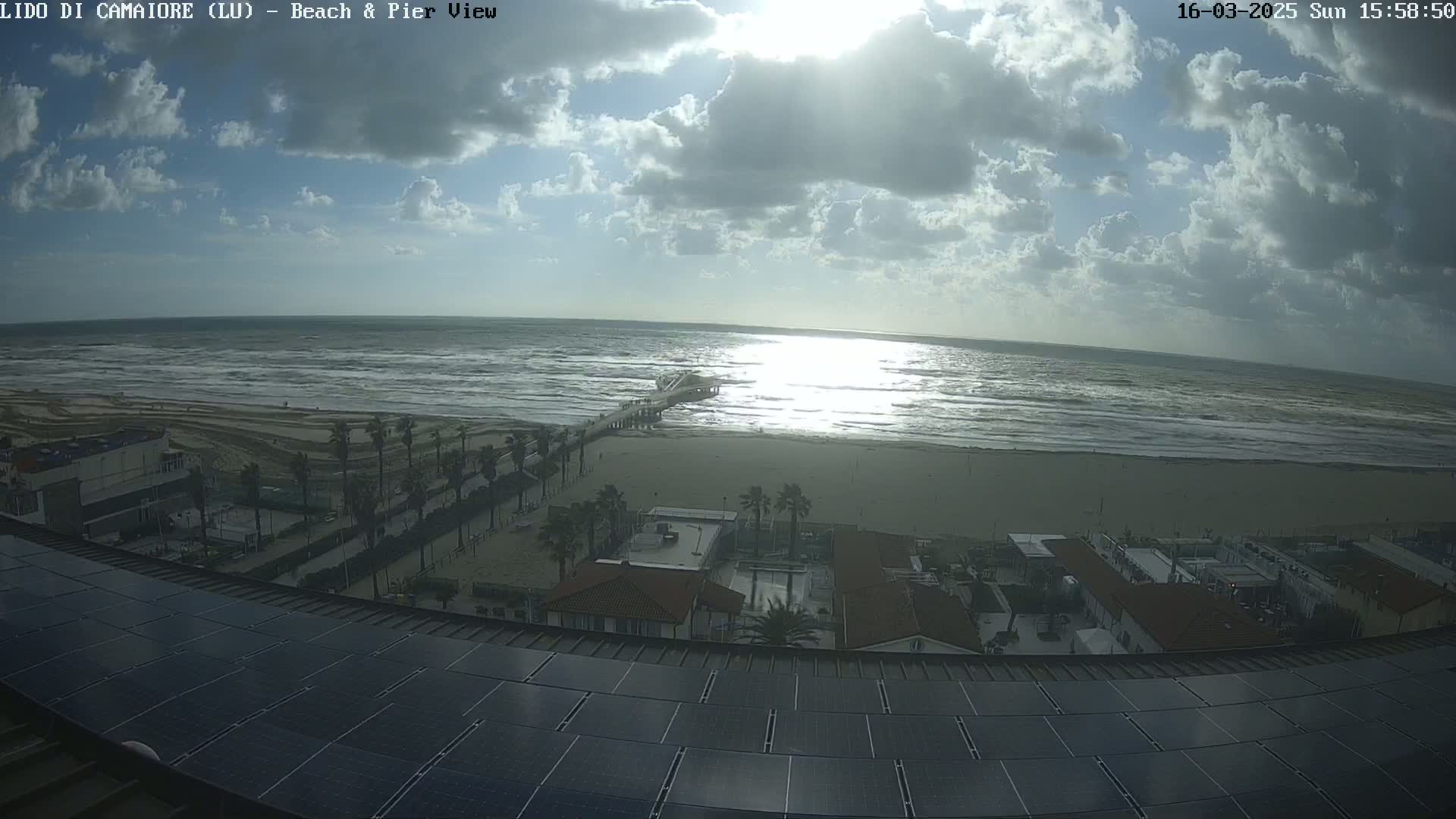 A partly cloudy day shows a beach with a pier, buildings, and palm trees, viewed from a rooftop with solar panels.