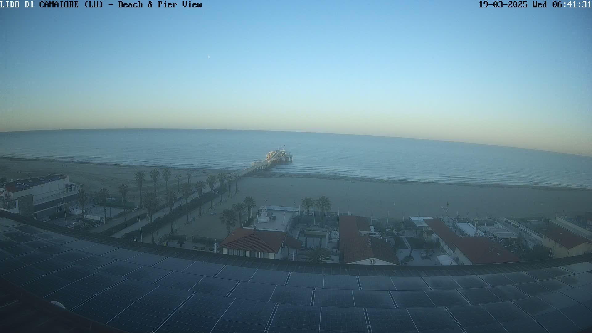 A high-angle view shows a beach with a pier extending into a calm, light-blue ocean under a clear, pale-blue sky.
