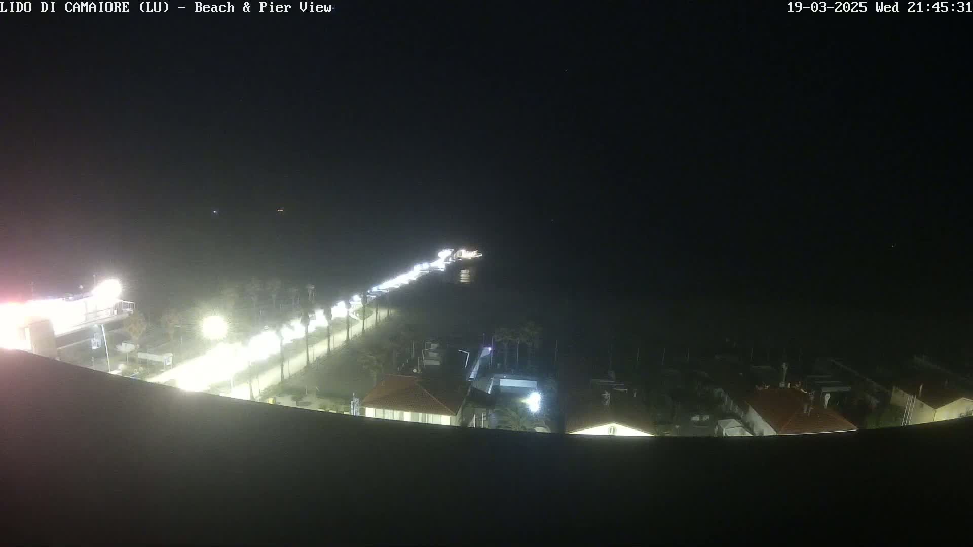 A nighttime, high-angle view of a beach and pier area, with a road lined with lights visible, and buildings illuminated below.