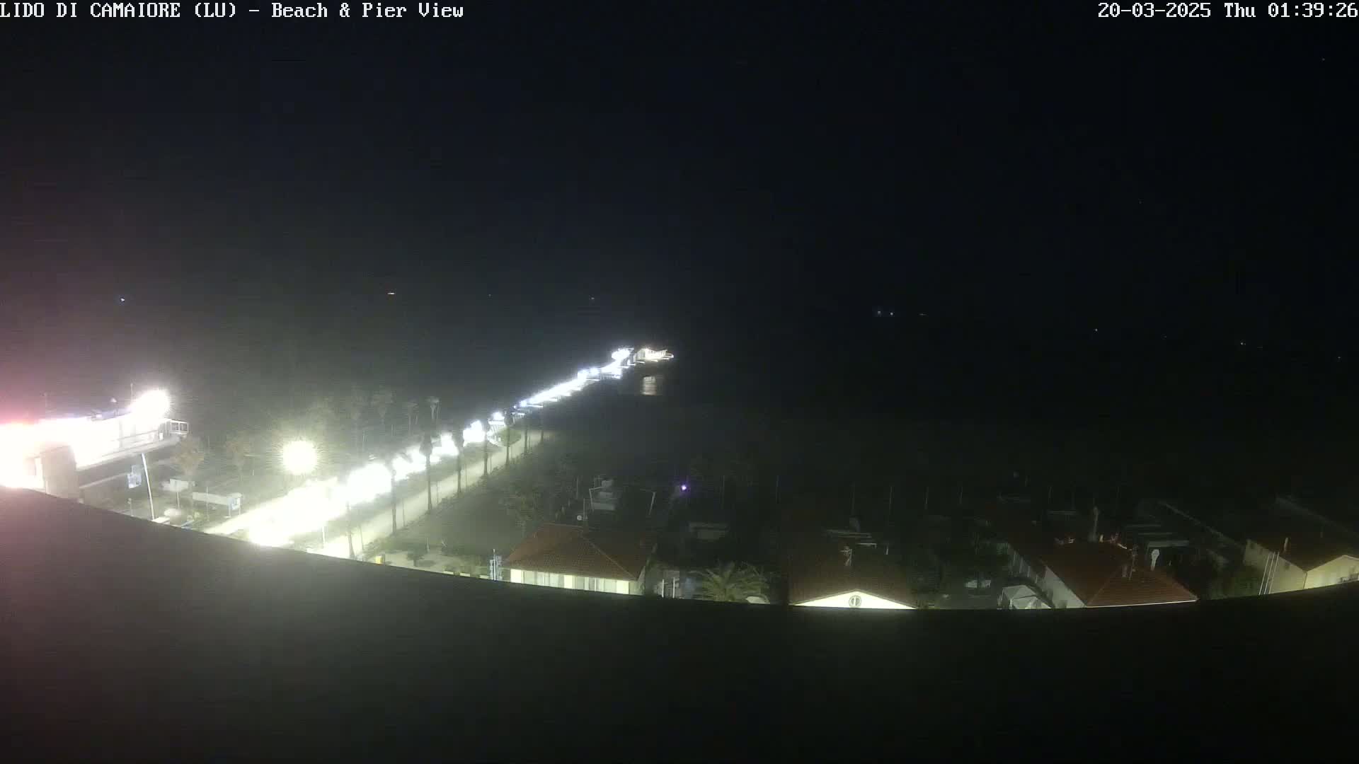 A nighttime, high-angle view shows a beach and pier area with dimly lit buildings and a road with cars visible.