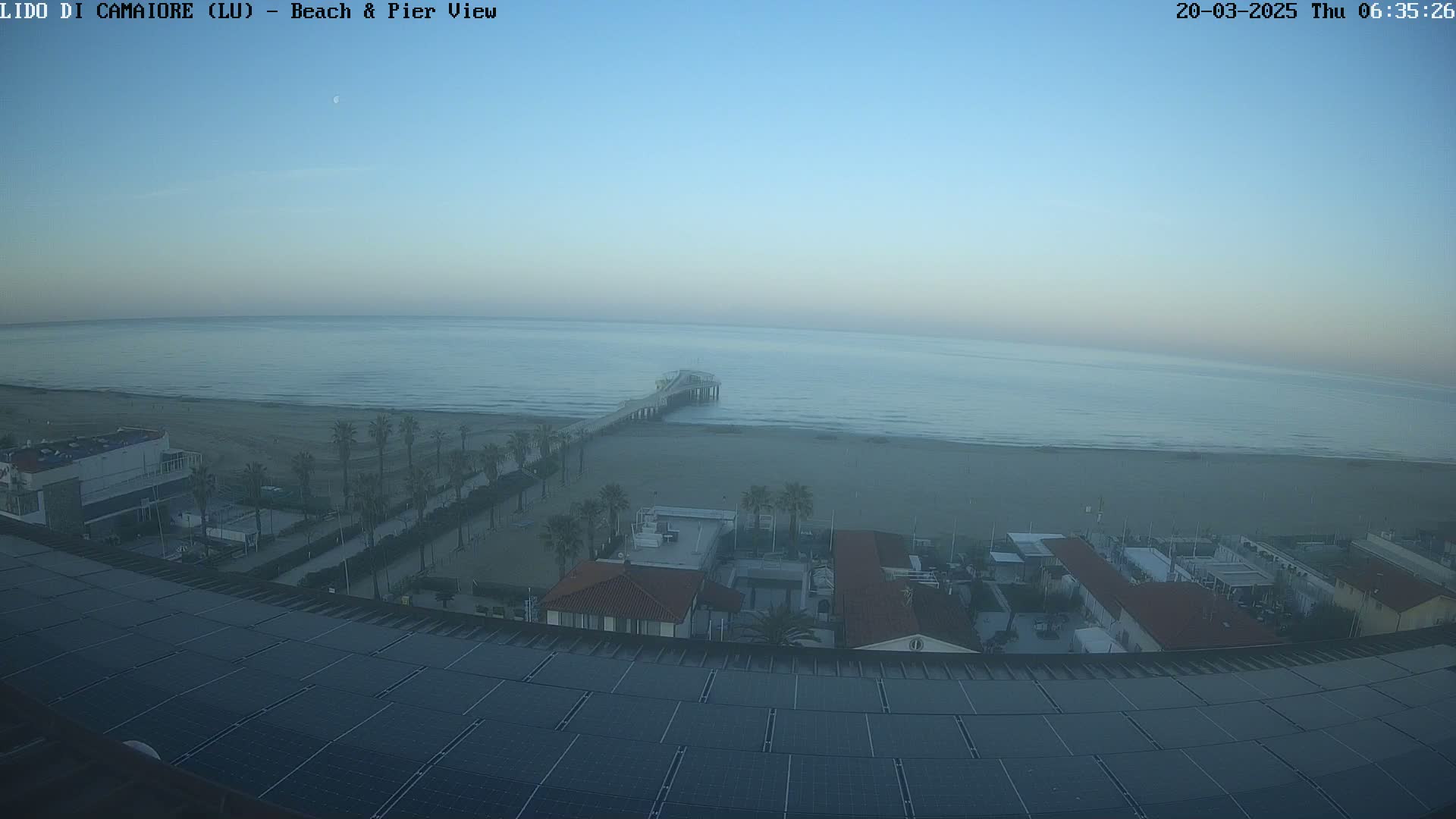 A calm ocean with a long pier extending into it, a sandy beach, and buildings with solar panels on a roof, all under a hazy, light-blue sky.