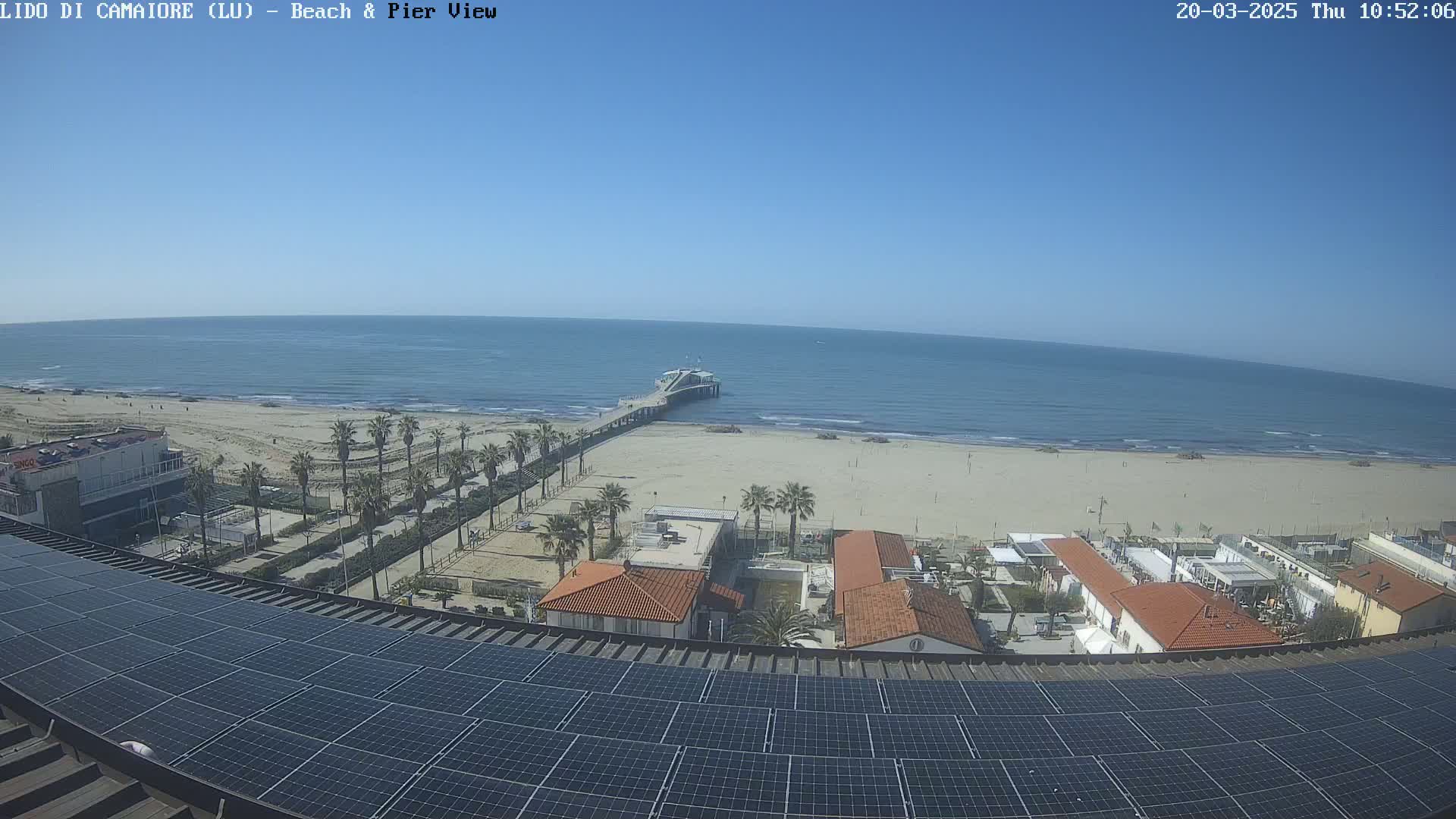 A mostly sunny beach scene with calm ocean waves, a pier, palm trees, and buildings is viewed from above a rooftop covered in solar panels.
