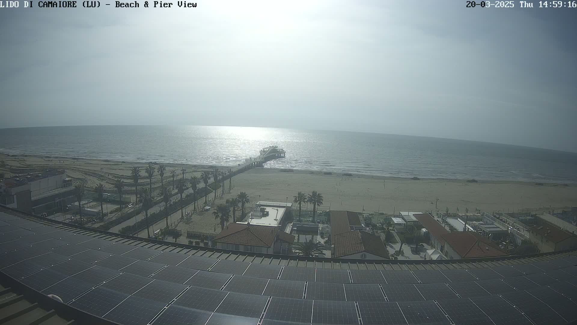 A pier extends into a calm, bright sea from a sandy beach, viewed from a rooftop covered in solar panels, with buildings and palm trees in the foreground.