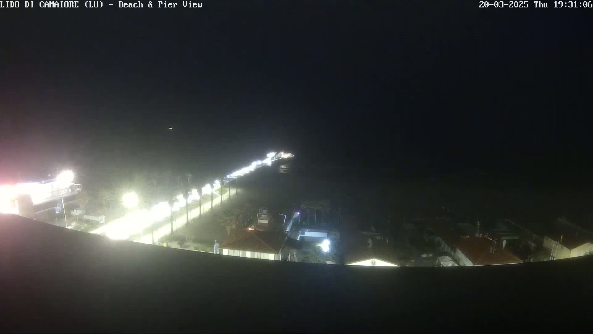 A nighttime, high-angle view shows a beach and pier area with illuminated buildings and a roadway lined with palm trees.