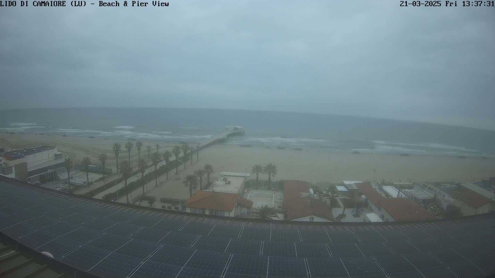 A beach scene on an overcast day shows a pier extending into a somewhat choppy sea, with palm trees lining a walkway leading from buildings to the sandy shore.