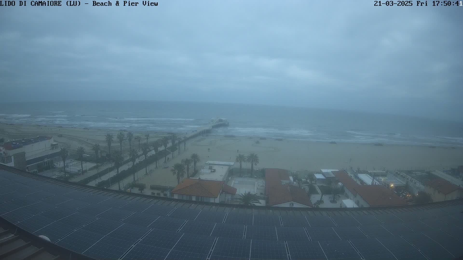 A seaside view on an overcast day shows a beach, pier, and buildings with solar panels on a roof, all under a hazy sky.