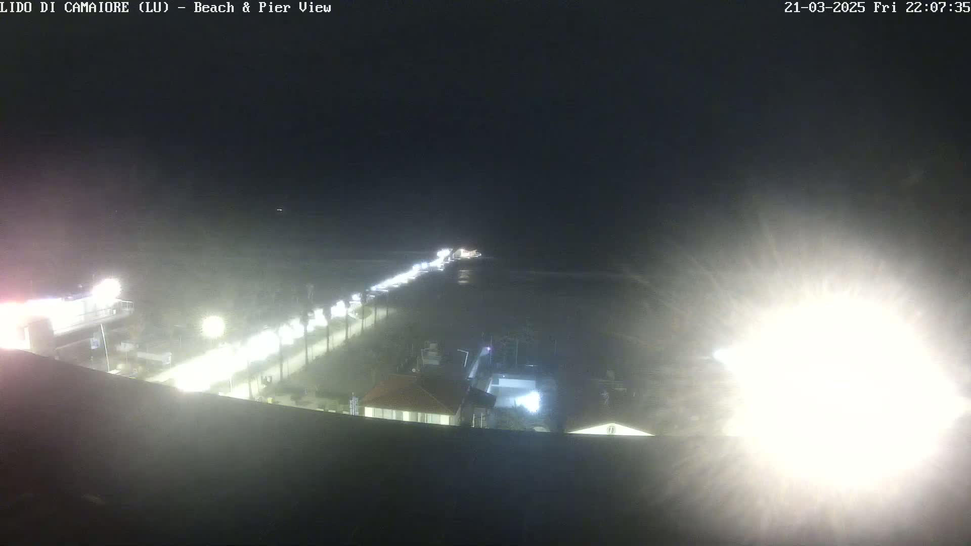 A nighttime, hazy view of a pier extending from a lit beachfront area into a dark ocean.
