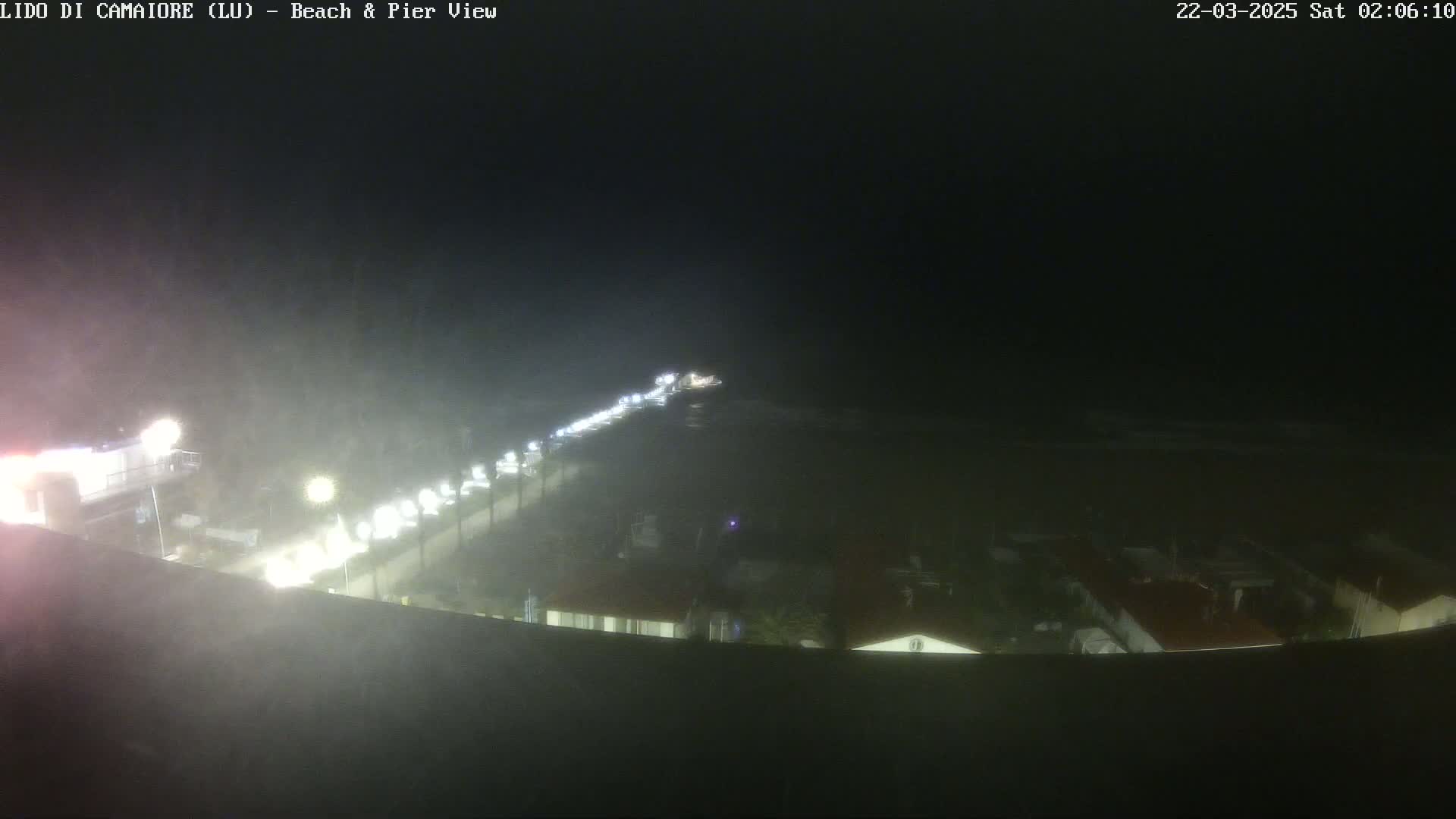 A nighttime, hazy view of a beach and pier area with lights visible along a road and buildings.