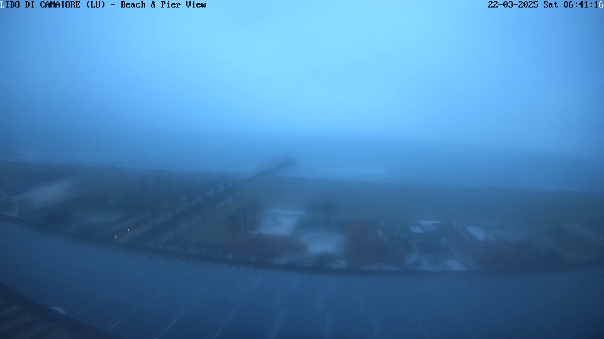 A hazy, blue-tinged view of a beach and pier, obscured by heavy fog or mist.