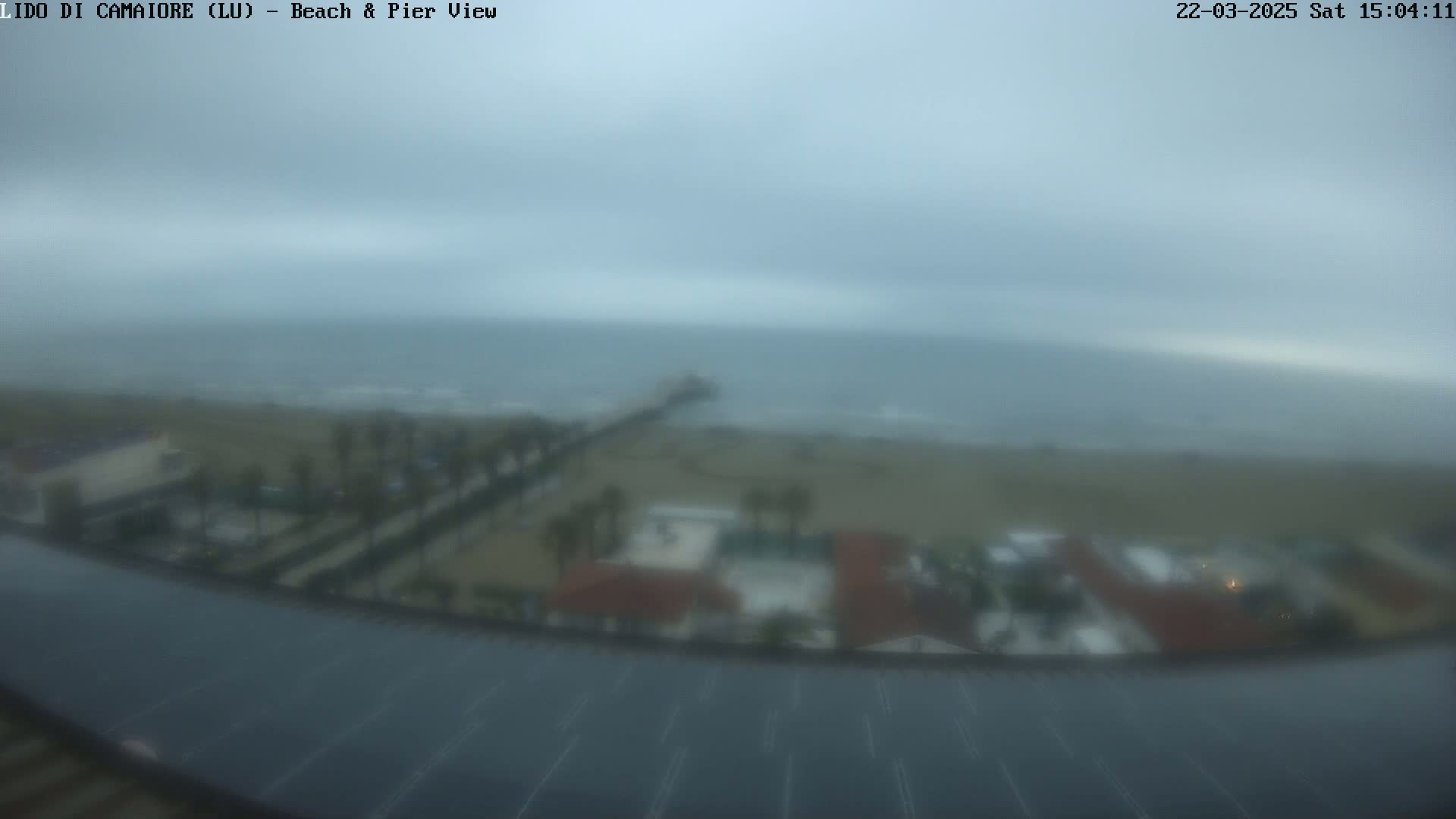 A hazy, overcast view shows a beach, pier, and coastal buildings from an elevated position.