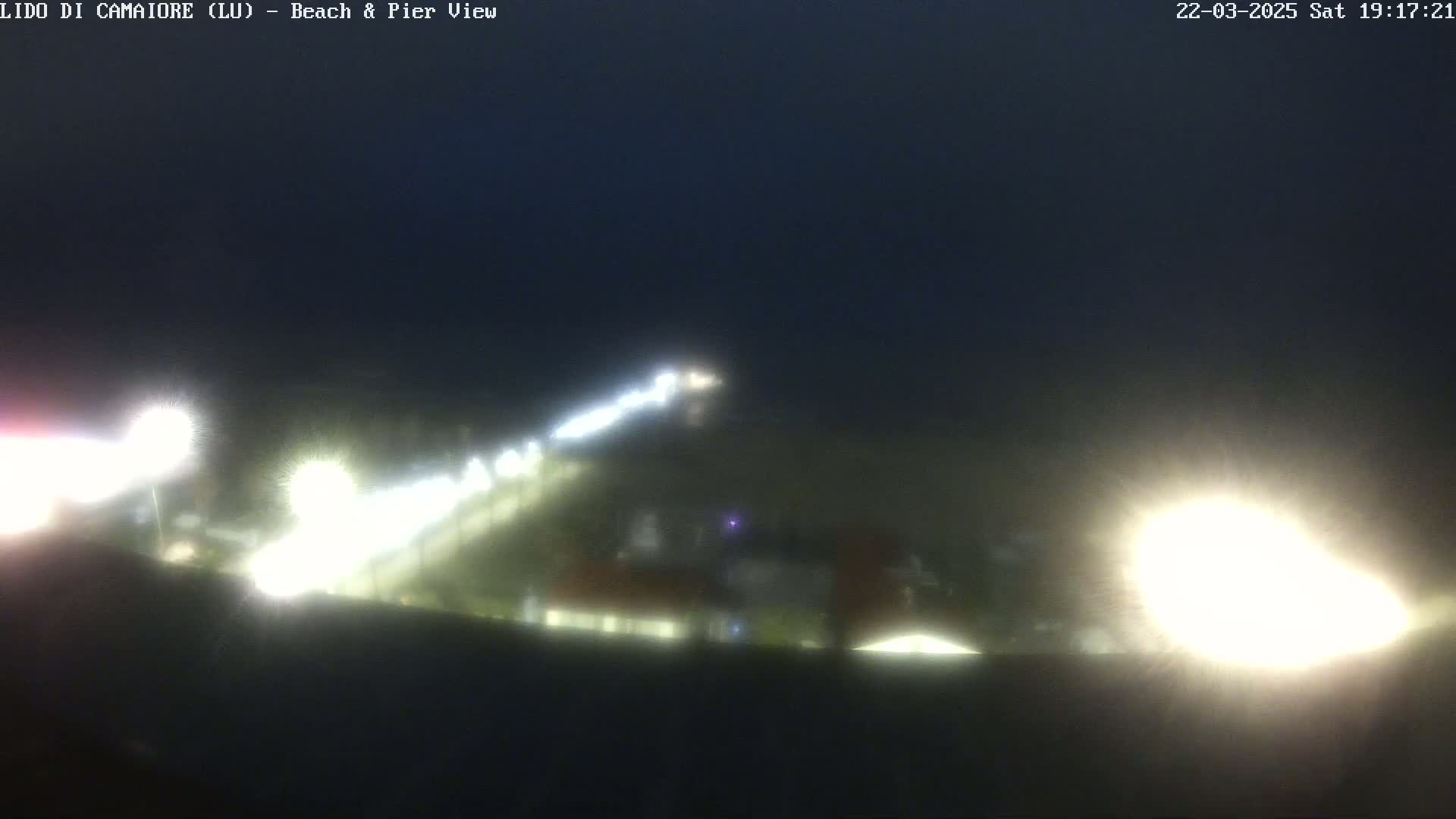 A blurry nighttime view of a beach and pier, illuminated by bright lights under a dark sky.
