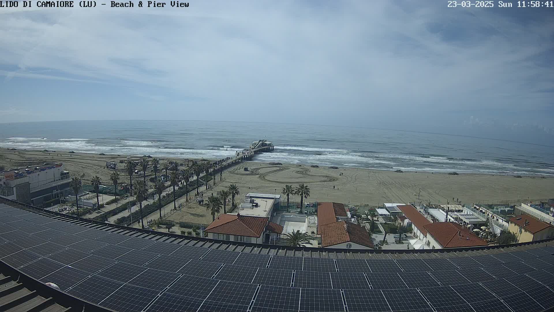 A mostly sunny day shows a beach with a pier, buildings, and palm trees, all viewed from above a large solar panel array.
