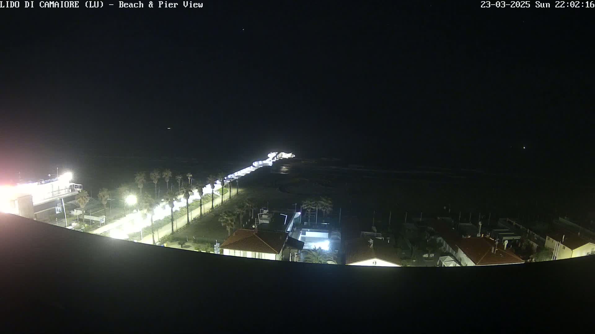 A nighttime aerial view shows a beach, pier, and buildings illuminated along a coastline under a clear, dark sky.