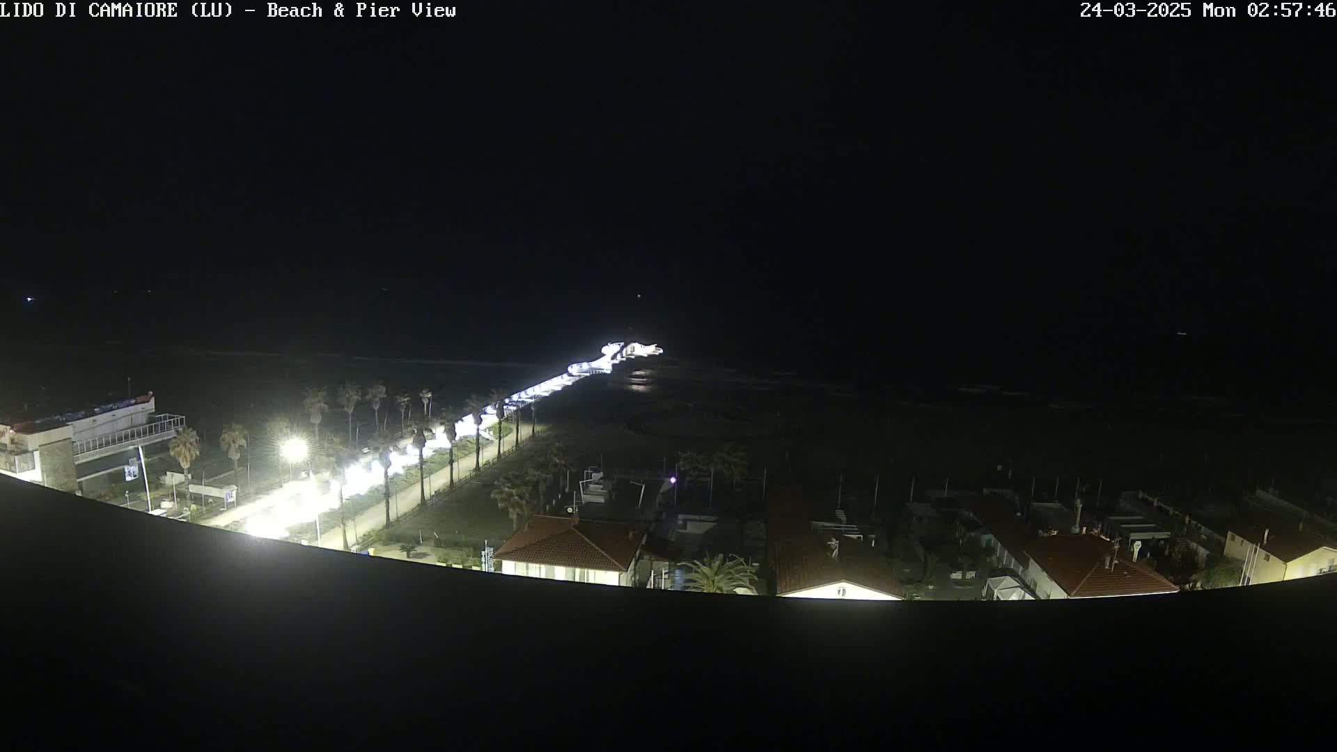 A nighttime, overhead view shows a beach, pier, and coastal road with buildings and palm trees, illuminated by artificial light.