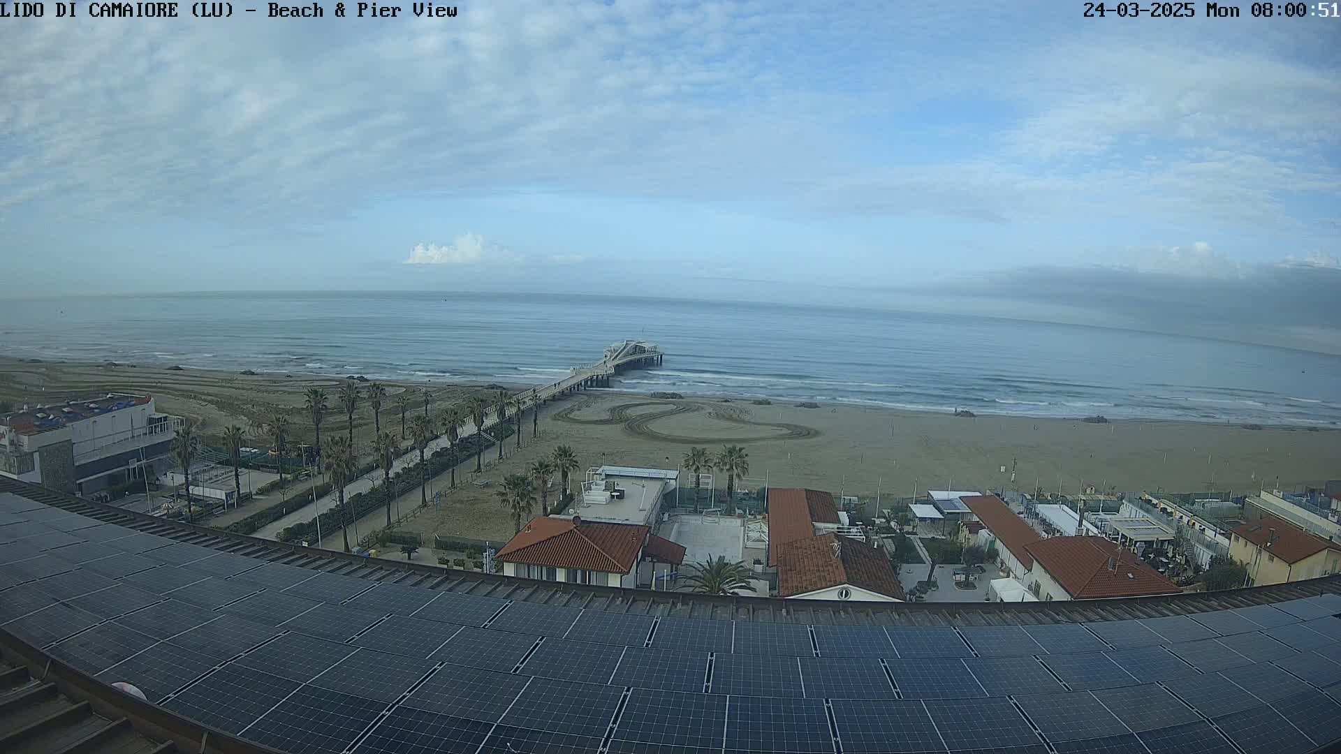 A mostly sunny day shows a beach with a pier, buildings, palm trees, and a large solar panel array on a rooftop overlooking the scene.