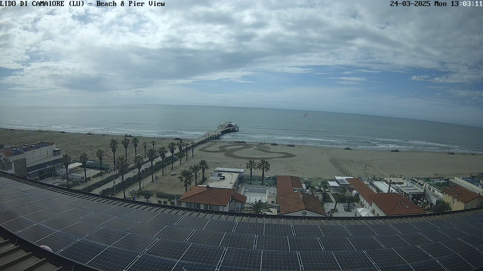 A mostly cloudy day shows a beach with a pier extending into the ocean, palm trees lining a paved walkway, and buildings with solar panels on the roof in the foreground.