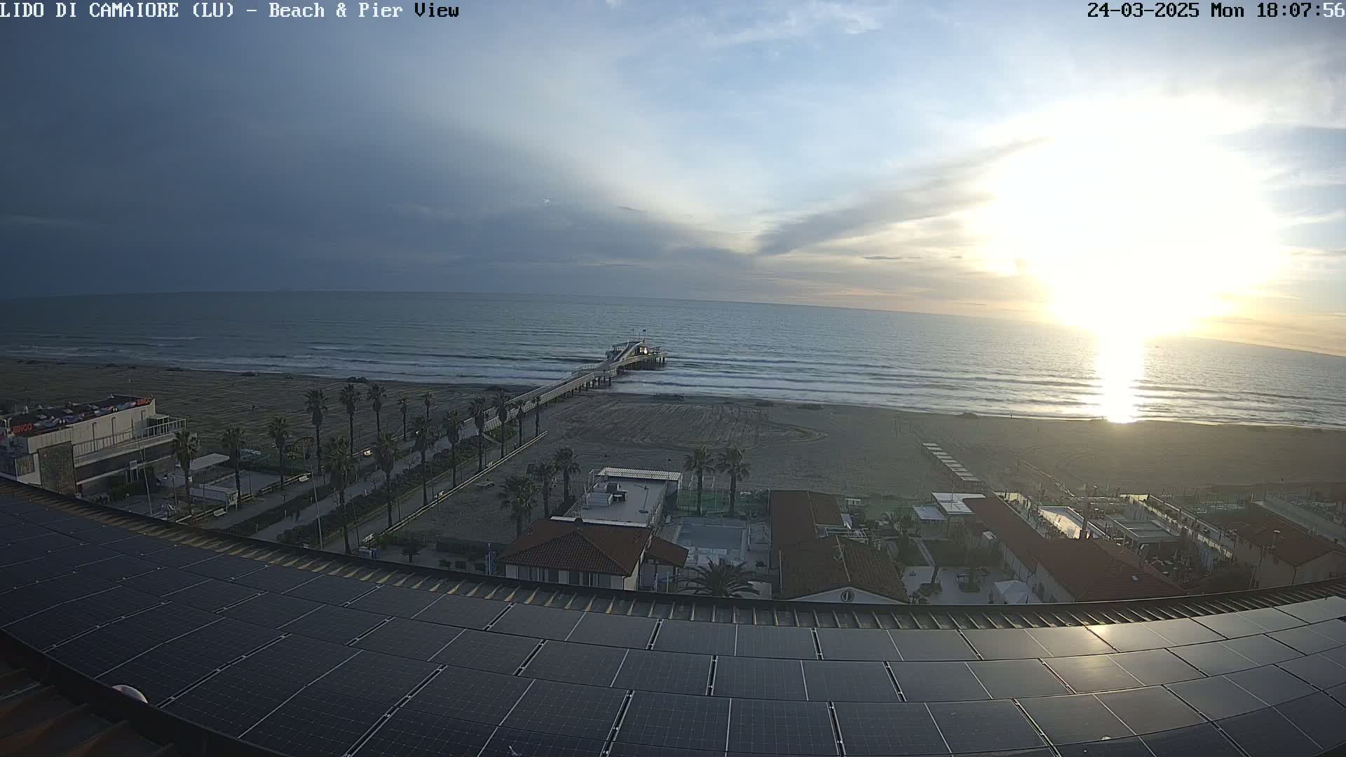 An aerial view shows a beach with a pier extending into a calm ocean under a partly cloudy sunset sky.