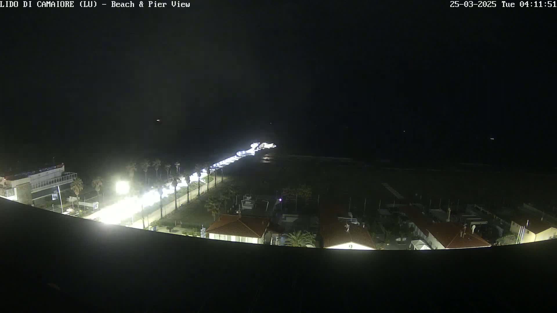 A nighttime, high-angle view shows a beach, a pier, and buildings along a coastline, illuminated by artificial lights.