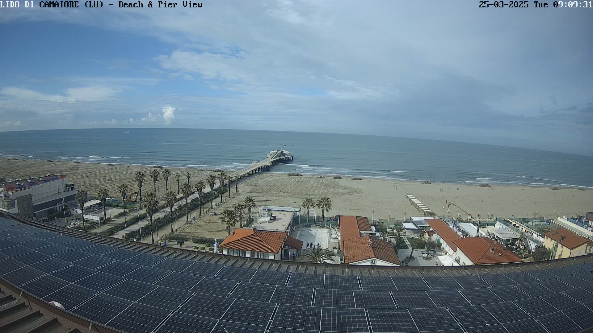 A mostly sunny day shows a beach with a pier extending into a calm sea, viewed from a rooftop covered in solar panels.