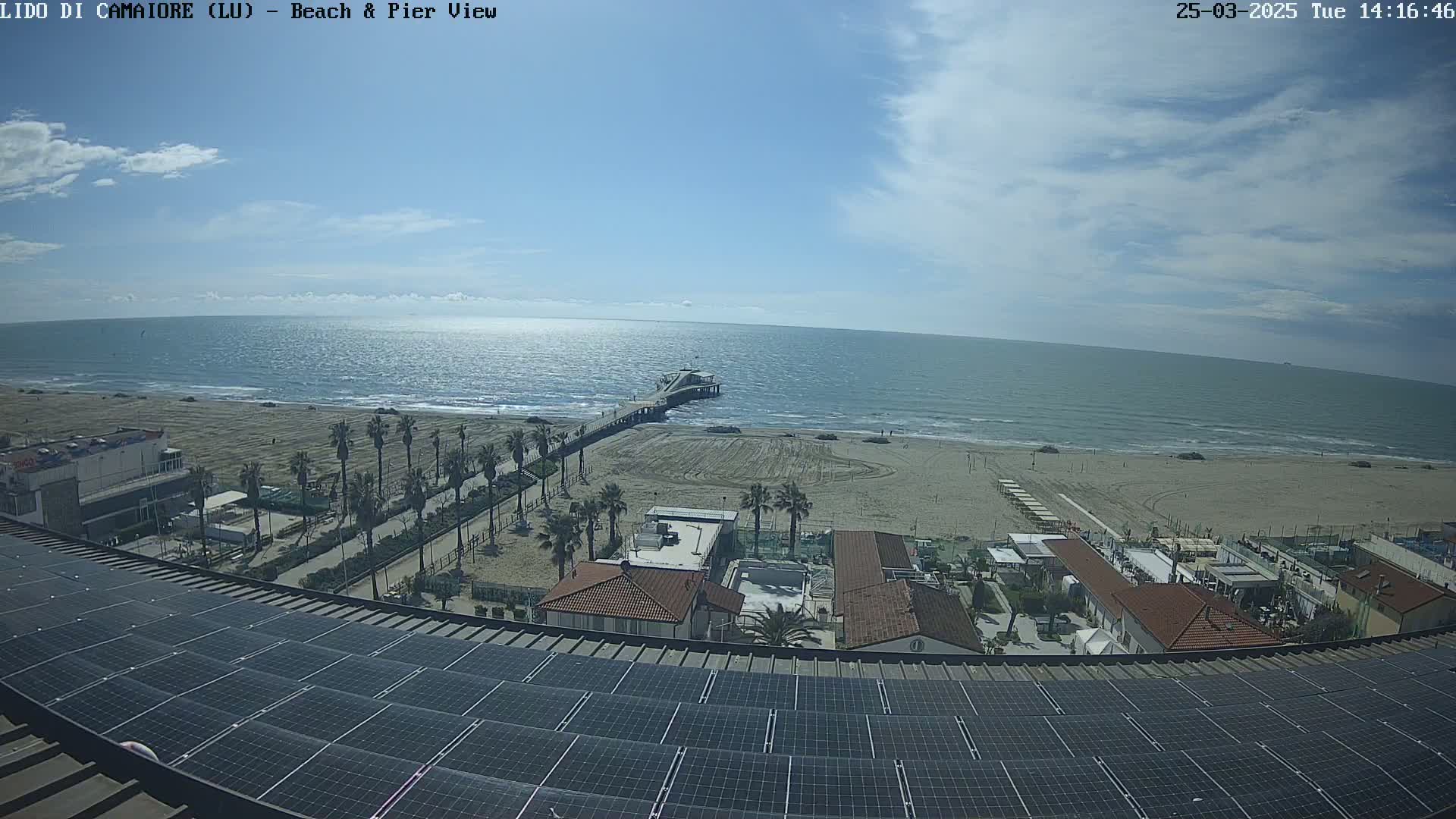 A mostly sunny day overlooks a sandy beach, ocean, pier, and palm trees from a rooftop covered in solar panels.